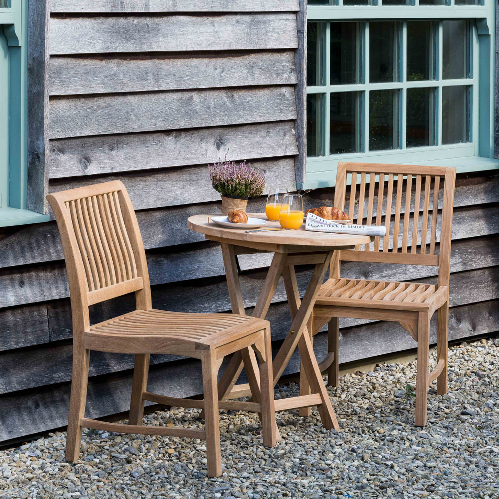 
                  
                    Teak round outdoor folding table with two glasses orange juice and pastries, and two teak dining chairs with slim slatted backrest and seats, on gravel floor, with wooden building in background
                  
                