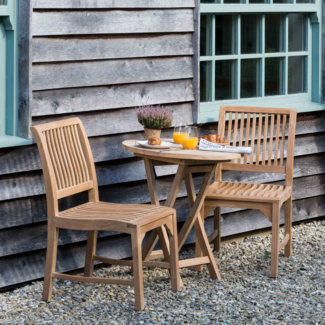Teak round outdoor folding table with two glasses orange juice and pastries, and two teak dining chairs with slim slatted backrest and seats, on gravel floor, with wooden building in background