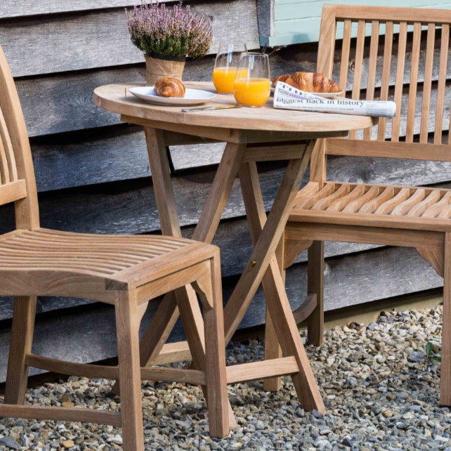Small round teak folding table set with orange juice and pastries, with two teak dining chairs, on gravel surface with wooden wall in the background