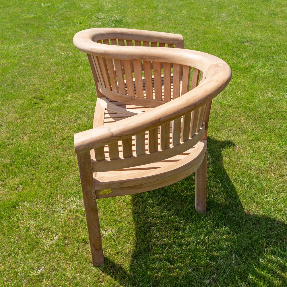 
                  
                    Teak love seat with curved, slatted, S-shaped backrest to seat two people next to each other but facing opposite directions, on sunny lawn. 
                  
                