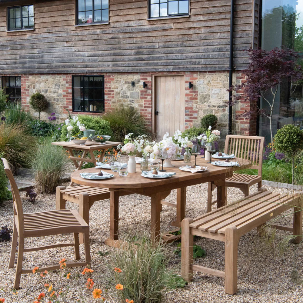 
                  
                    Teak benches without back or armrests, and teak dining chairs, around teak oval dining table, set with plates, glasses and flowers, with brick and wood building behind. 
                  
                