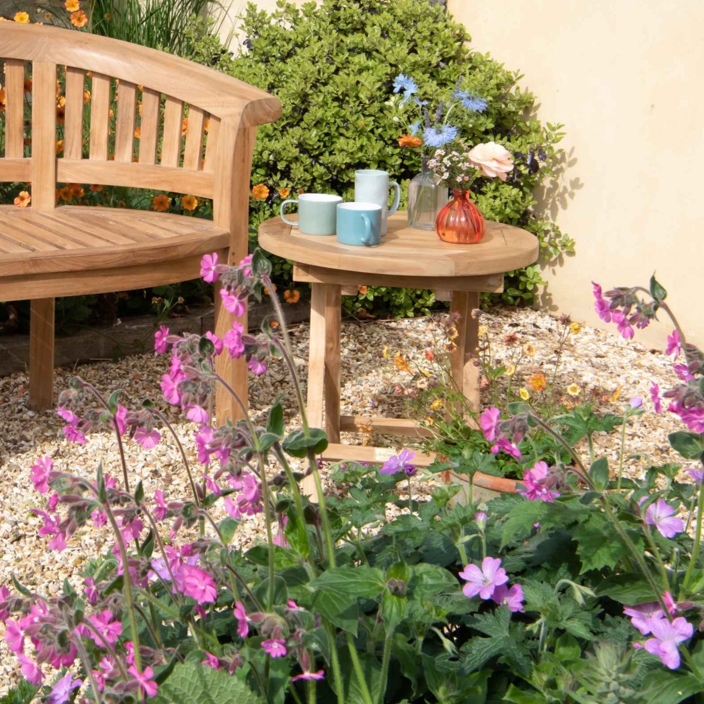 Teak Round Folding Coffee Table set with cups and flowers, next to a curved teak bench, on gravel patio with purple flowers in the foreground