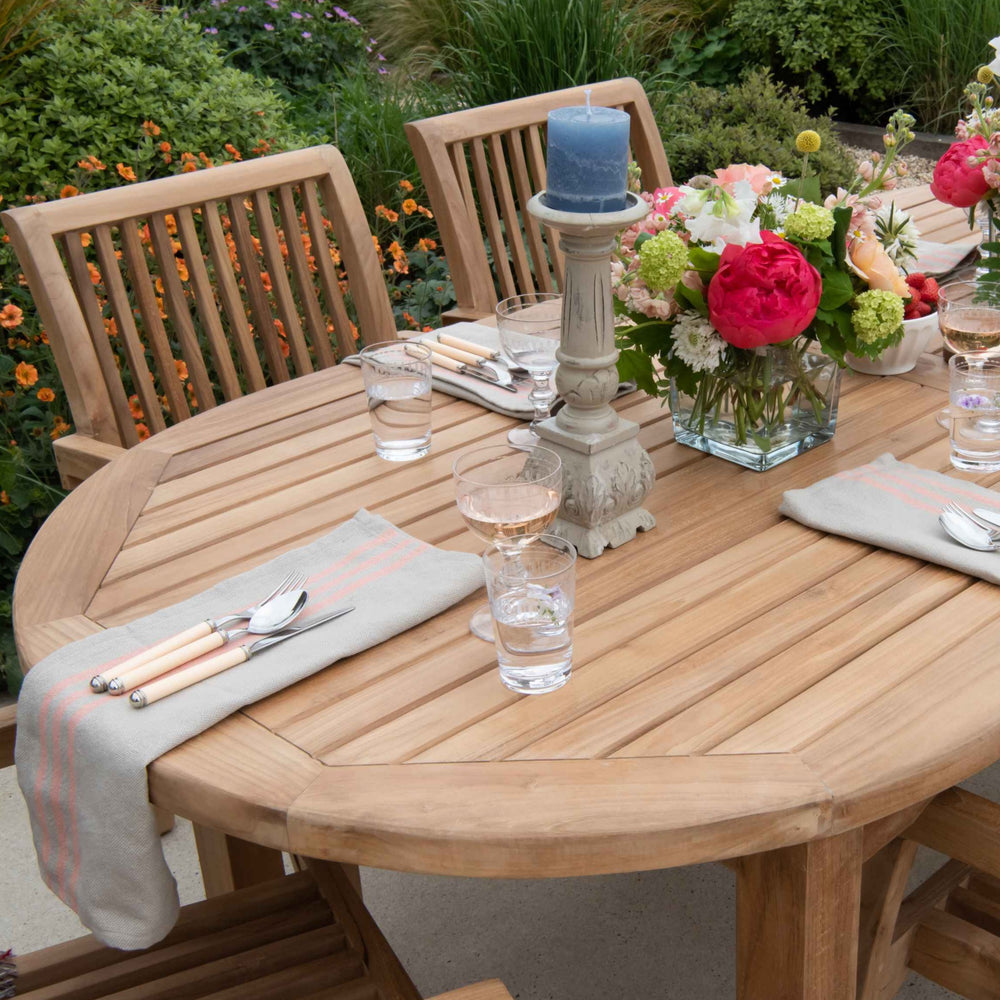 
                  
                    Teak oval dining table and chairs, laid up with flower arrangements, candlesticks and place settings in a garden setting. 
                  
                