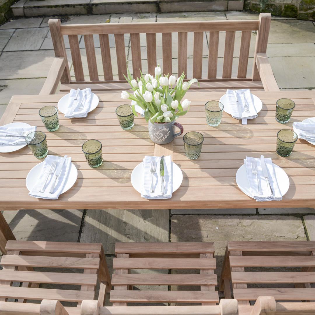 Teak rectangular dining table, set with seven place settings, a teak bench on one side and three teak dining chairs on the other, on stone patio. 