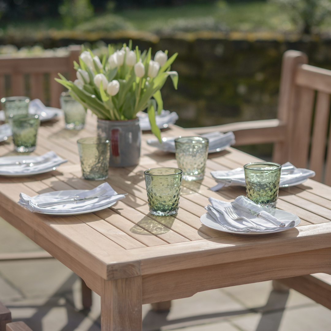 Rectangular Teak Dining table with place settings, glasses and flowers with teak dining chairs in background, in patio garden setting. 