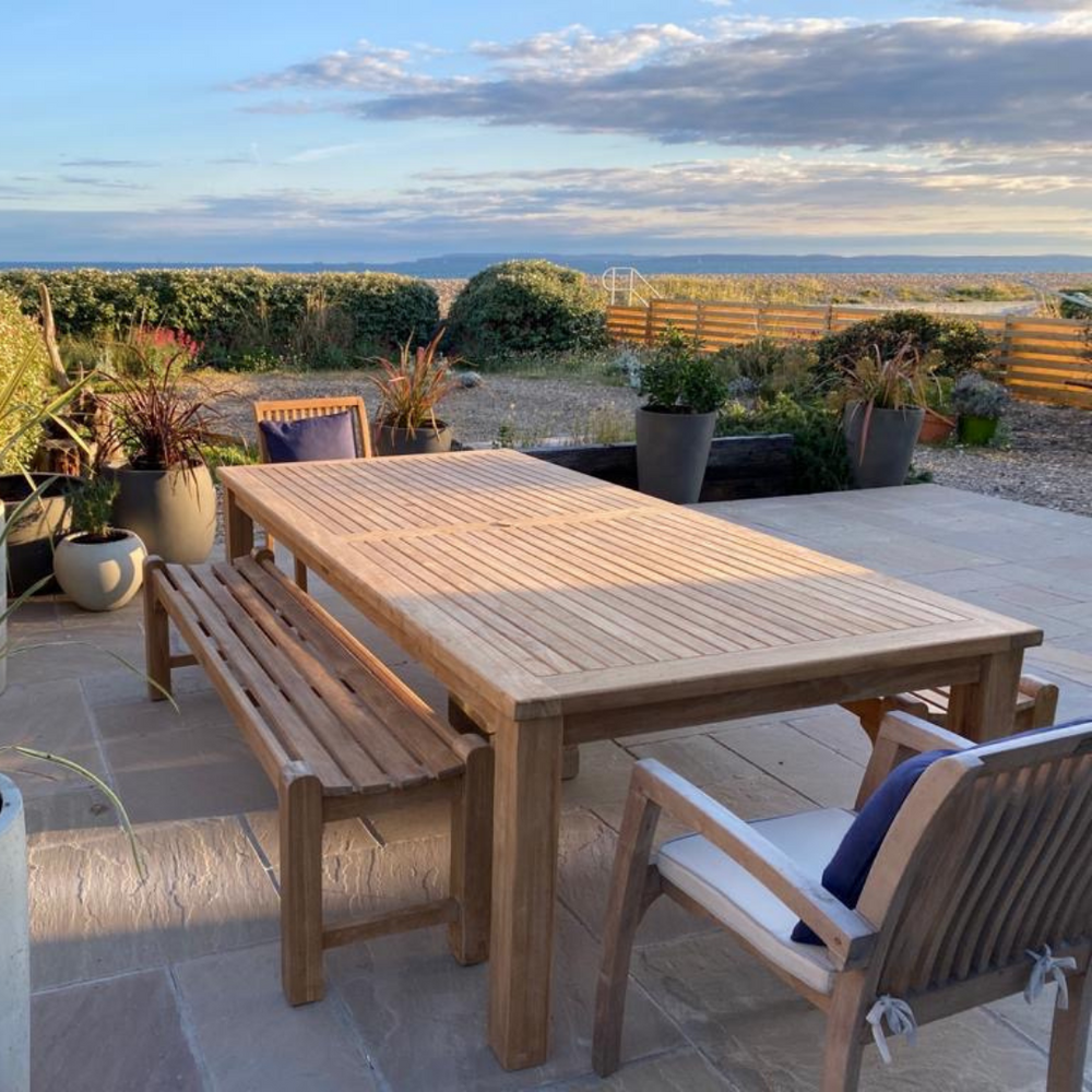 
                  
                    Teak rectangular dining tale with teak bench and dining chairs, on stone patio with planters, with far reaching views of dunes and hills.
                  
                
