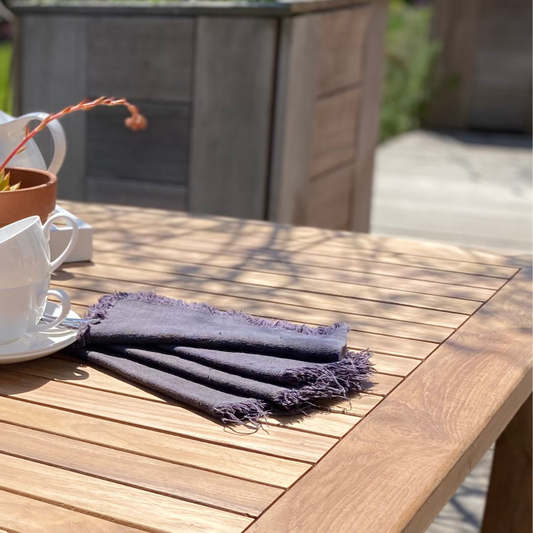 Close up of corner of square teak coffee table with napkins, cups and saucers, in garden setting. 