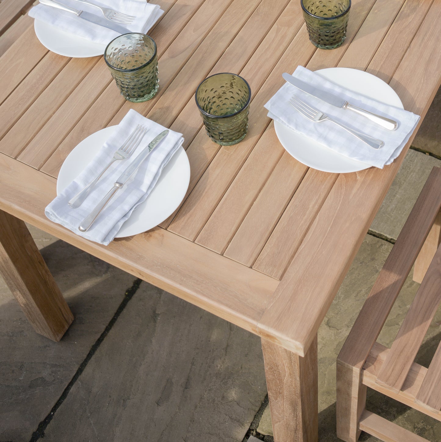 A set square teak table with a solid square frame and slatted tabletop, set outdoors with plates, glasses, and cutlery arranged for a meal.  