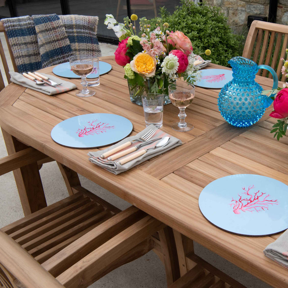 
                  
                    Teak oval dining table with teak dining chairs set around. On the table are blue placemats, blue glass jug, water and wine glasses and vases with flowers. 
                  
                