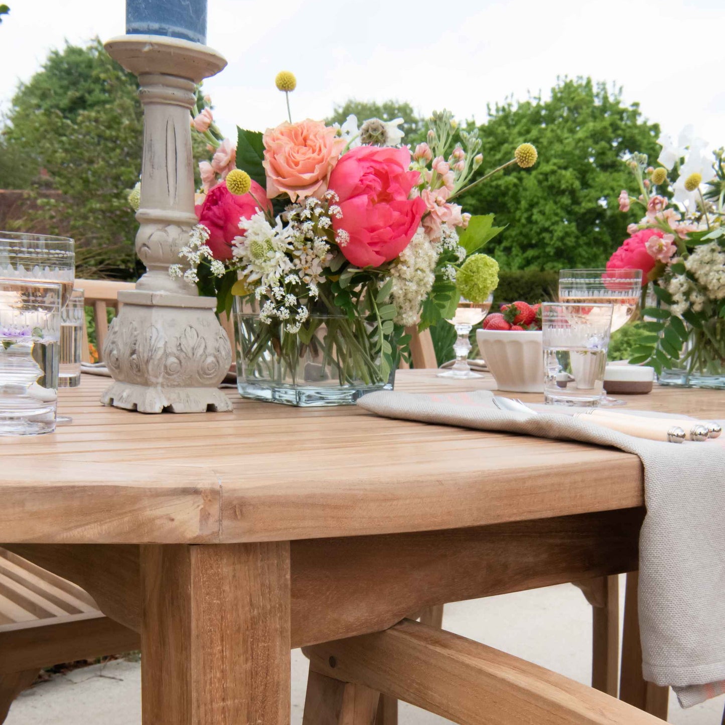 Close up of edge of teak oval outdoor dining table with flower arrangements, candlesticks and place settings. 