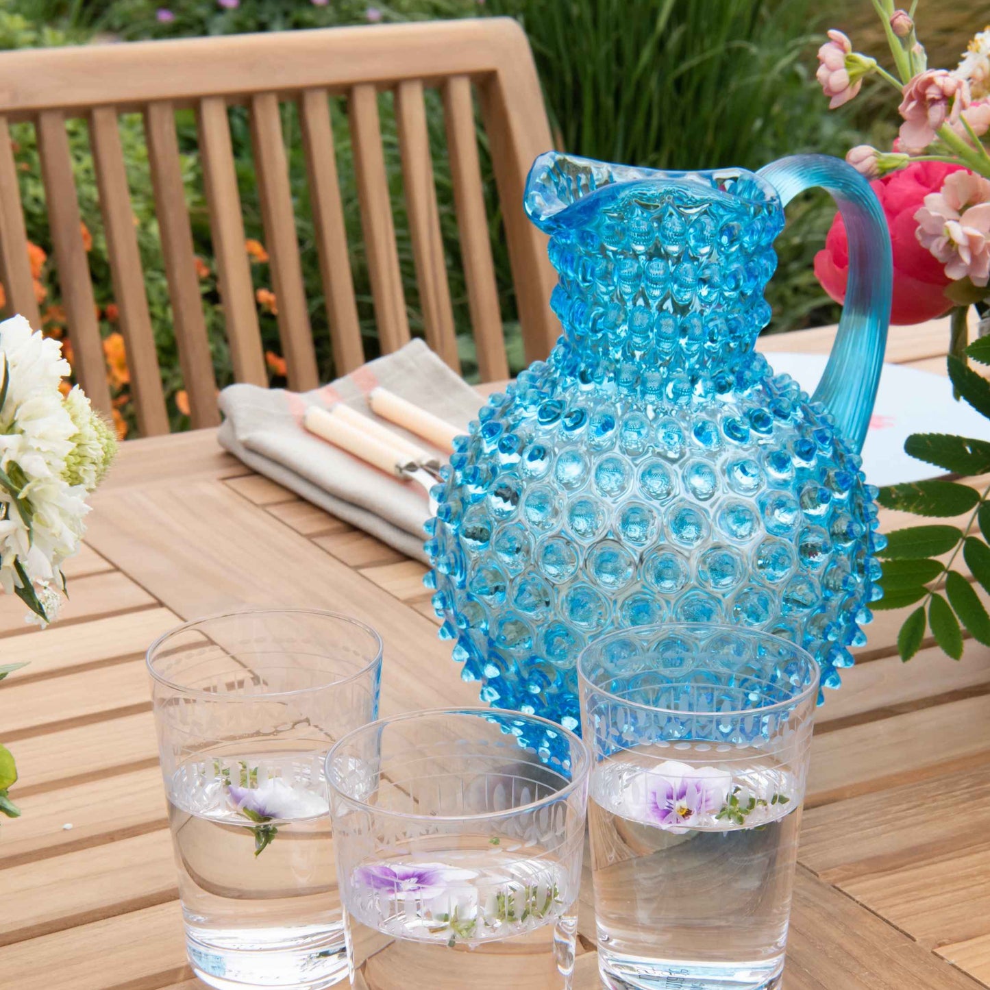 Teak oval dining table set with a vase of flowers, a blue glass jug and clear water glasses, with a teak dining chair and place setting.  