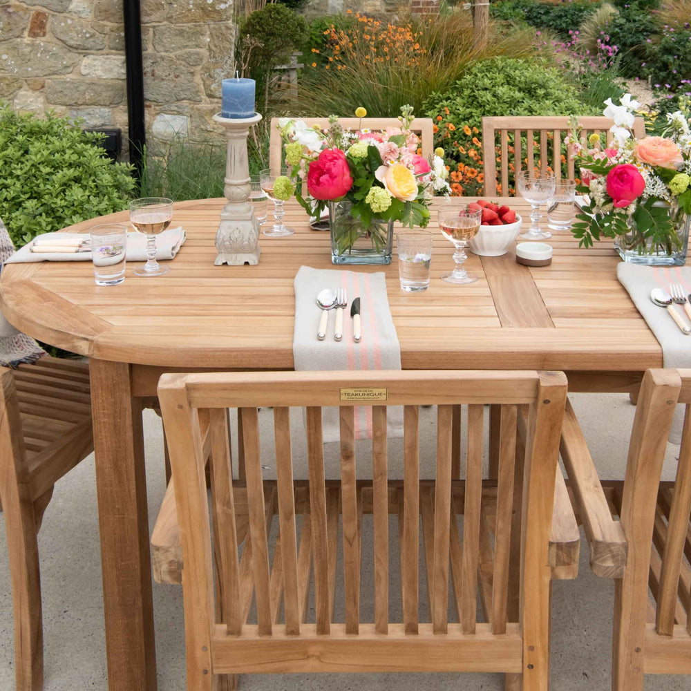 
                  
                    Teak oval dining table and chairs, set with vases of flowers, place settings and candlestick, with flower beds behind. 
                  
                