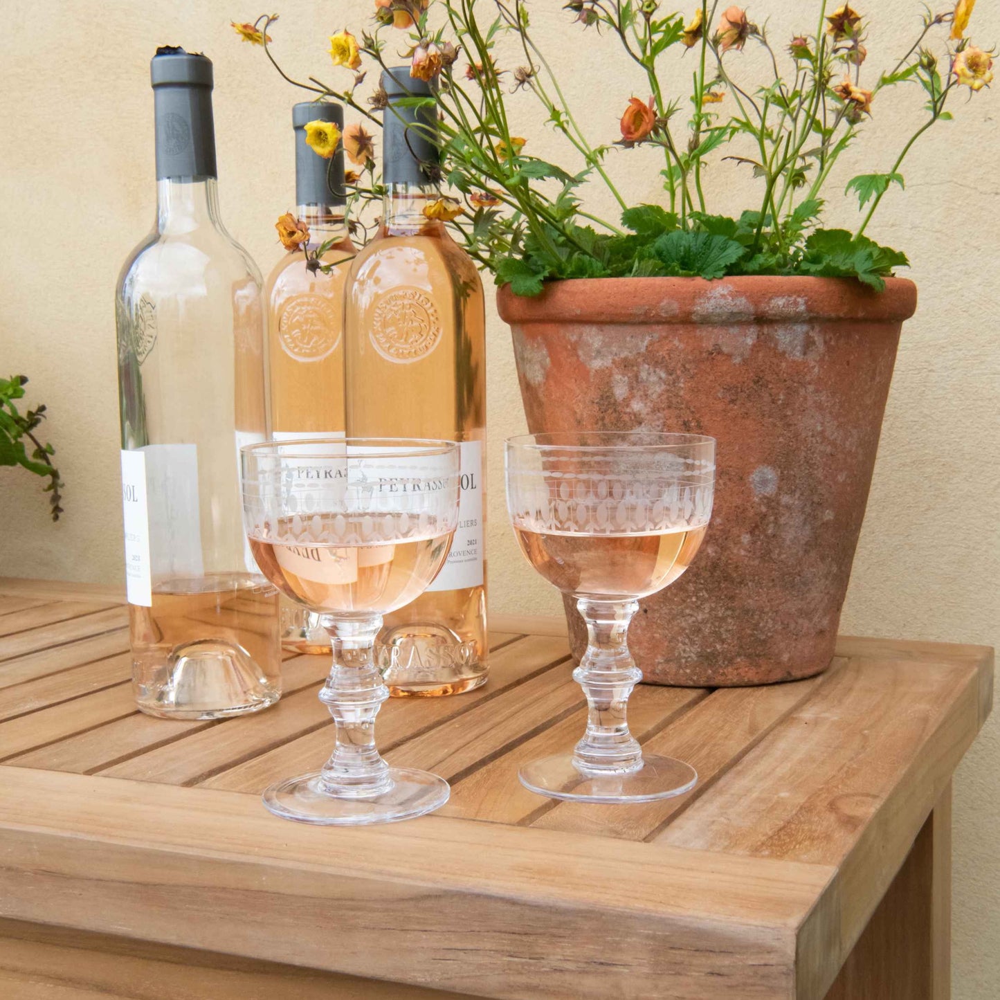 Teak rectangular side table close up with two full wine glasses and wine bottles behind, next to terracotta plant pot.  