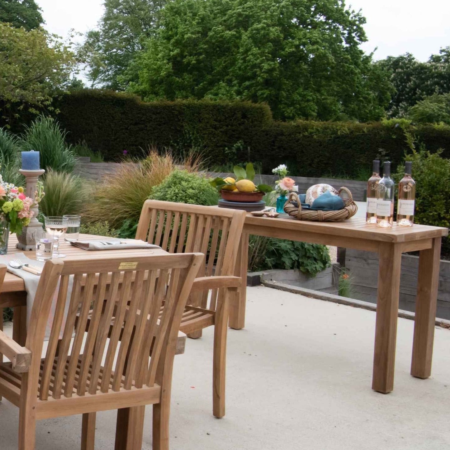 Teak Rectangular Side Table with wine, fruit and decorative items, on a stone patio next to teak outdoor dining table and chairs, with garden behind.  