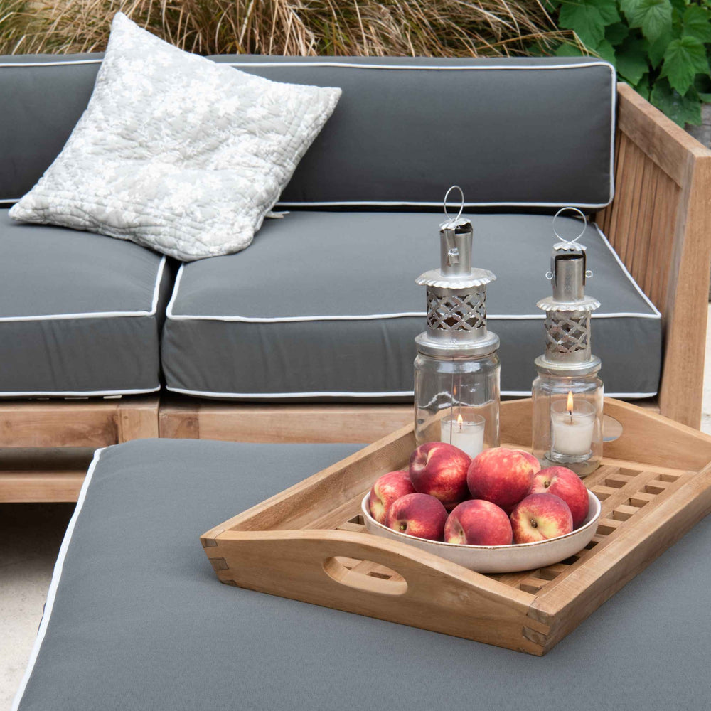 
                  
                    Teak modular corner outdoor sofa with teak ottoman stool, all with grey and white cushions. The stool is holding a teak tray with apples and lanterns. 
                  
                