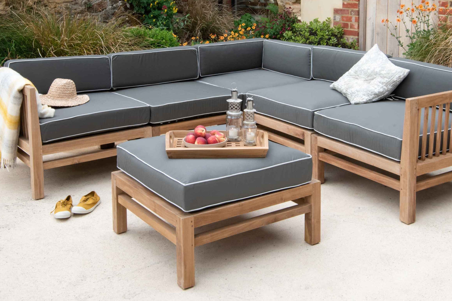 Teak Modular Corner Outdoor Sofa with green - grey seat and back cushions. In front is teak ottoman with same coloured cushion and tray of candles and fruit.  