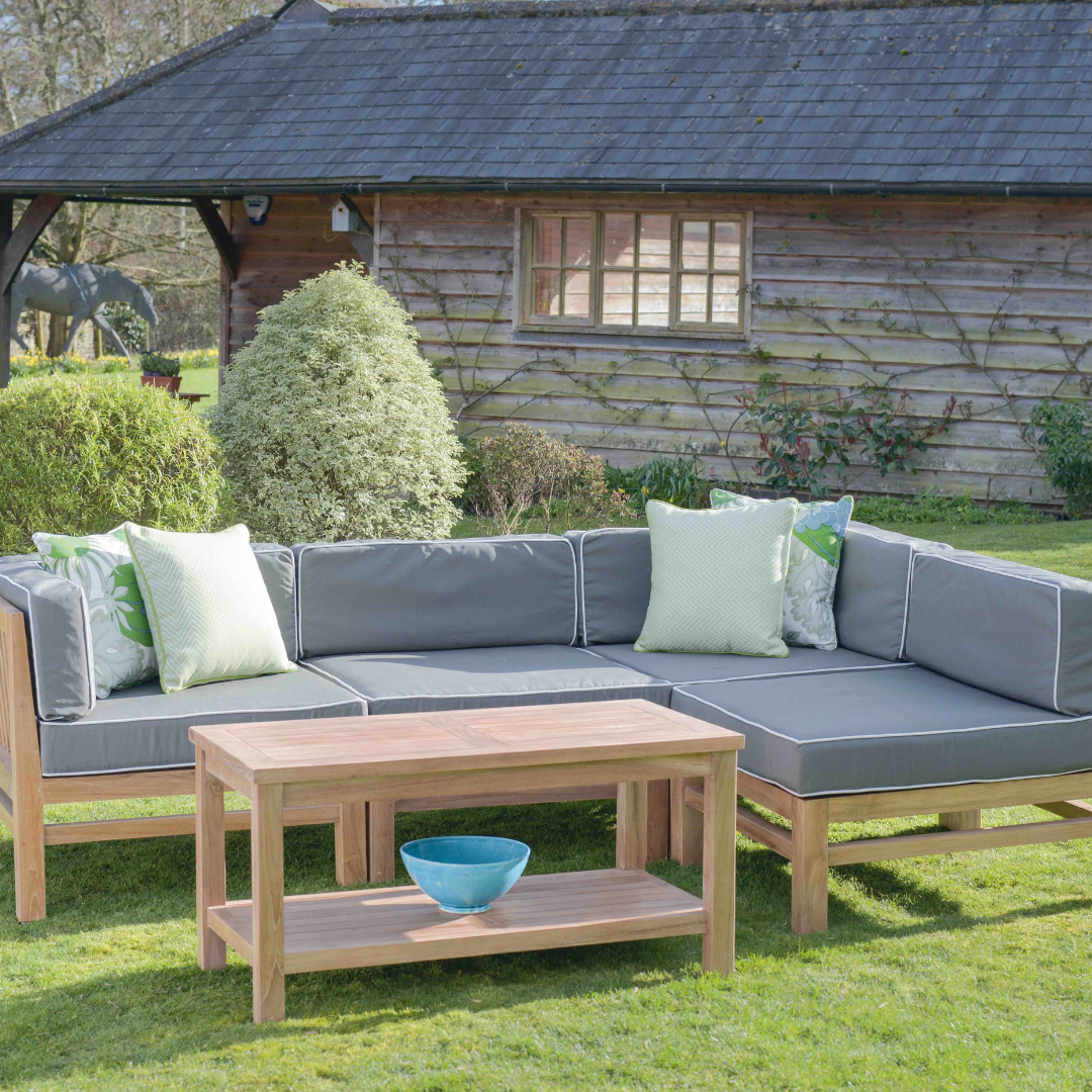 Teak modular corner outdoor sofa with green-grey seat and backrest cushions, paler patterned scatter cushions, and teak coffee table with shelf, in garden in front of wooden building. 