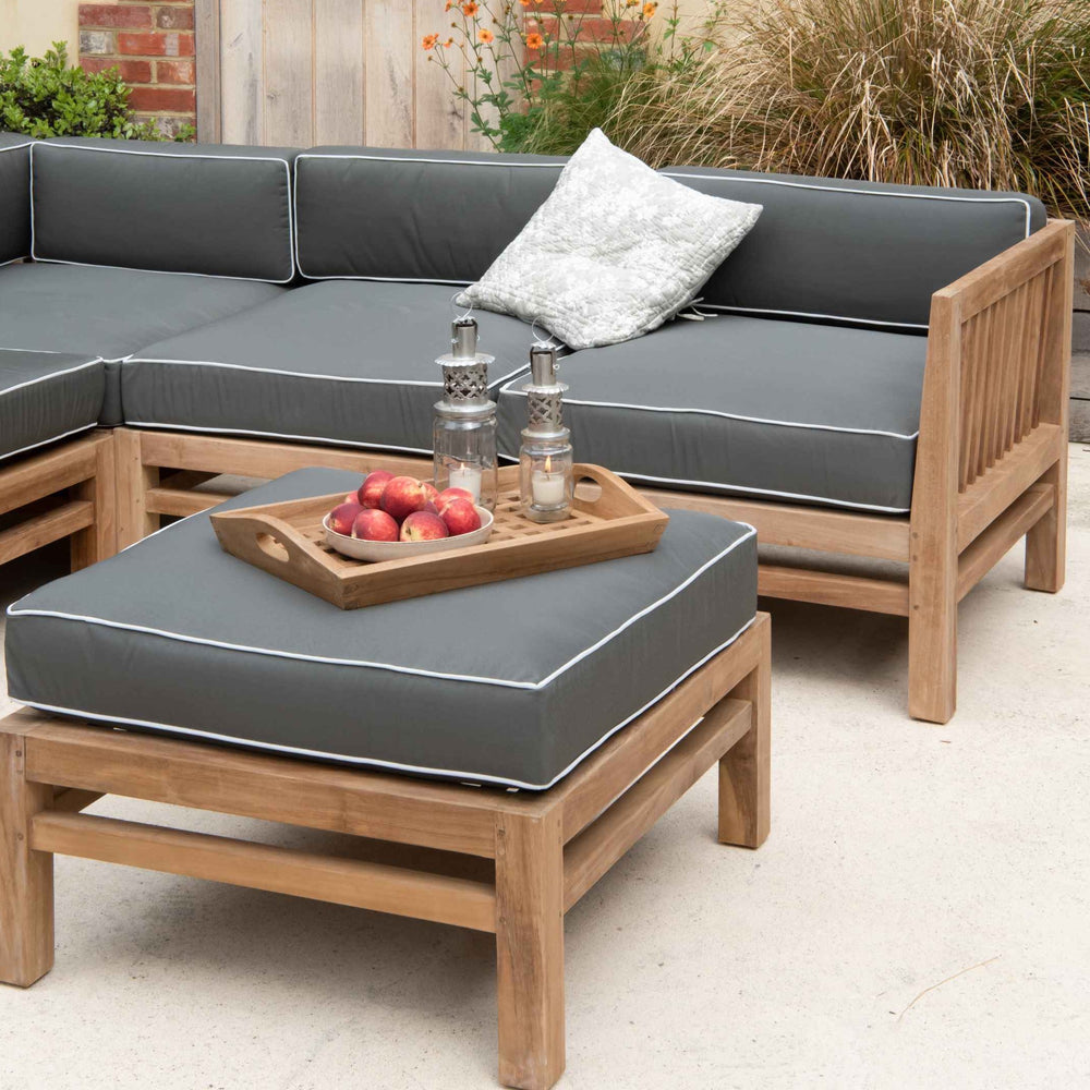 
                  
                    Teak Outdoor Sofa and ottoman stool with grey and white cushions on a stone patio. On the stool is a teak tray with fruit and lanterns.  
                  
                