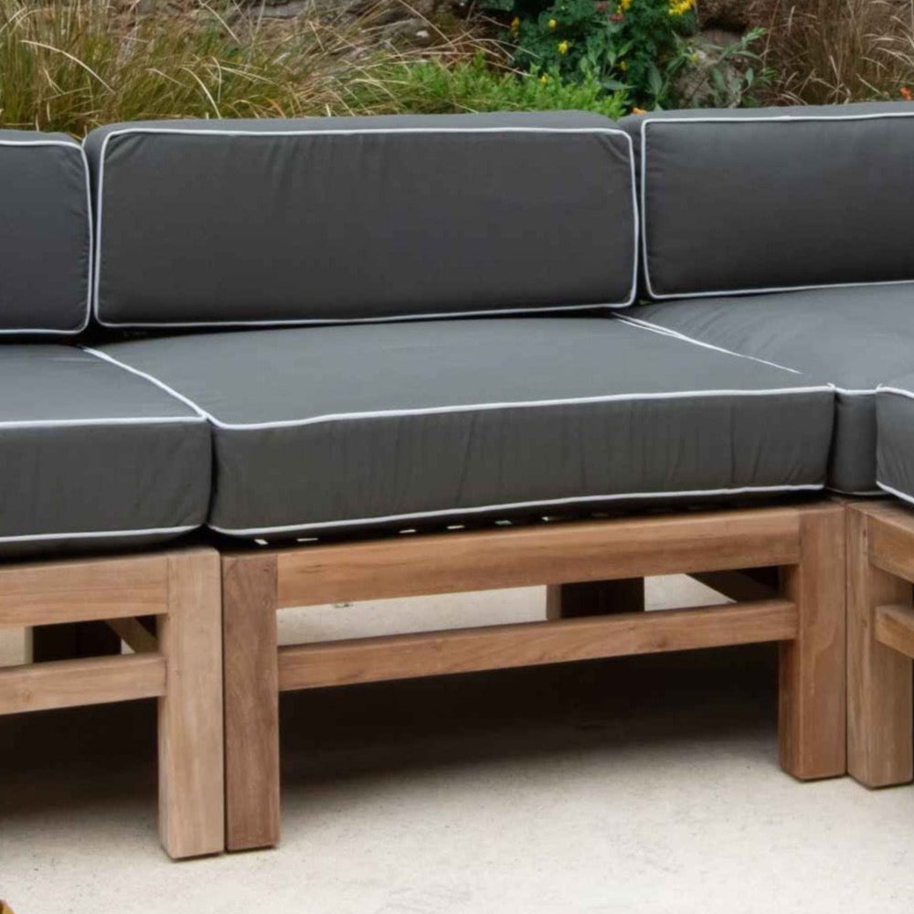 
                  
                    Mid-section of teak outdoor sofa with dark grey and white cushions, on stone surface with greenery behind
                  
                