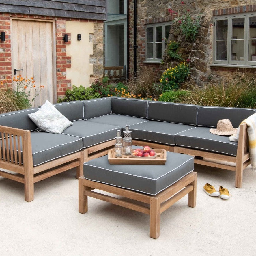 Teak outdoor corner sofa and ottoman stool all with dark green and white cushions, with a tray of fruit, a straw hat and pair of shoes around, in front of brick building 