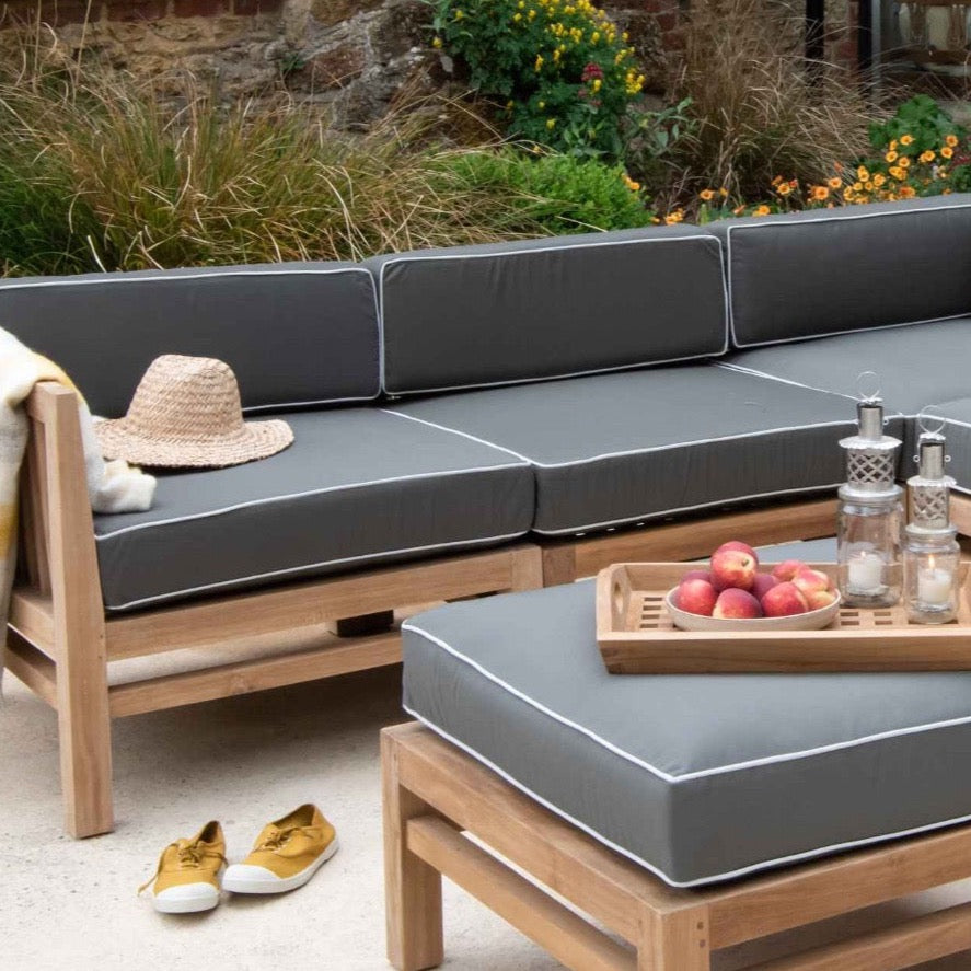 Teak outdoor sofa with green and white cushions, holding straw hat. In front is a teak ottoman stool holding tray of fruit and lanterns. 