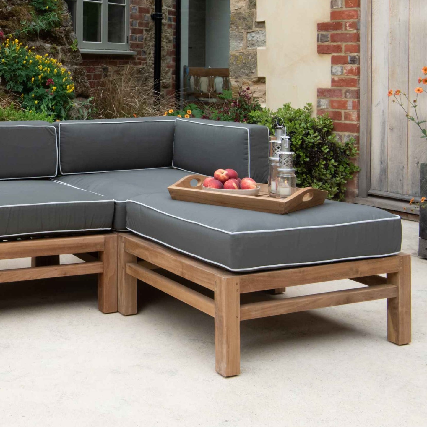 Teak outdoor sofa and teak ottoman stool with green and white cushions, holding teak tray of fruit and lanterns, on stone patio in front of brick building
