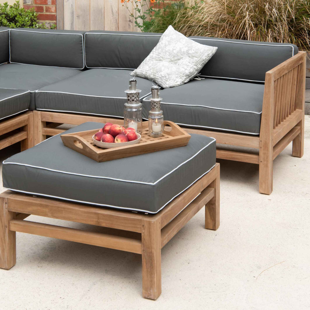 
                  
                    Teak outdoor sofa and ottoman stool with dark grey and white cushions, on a stone patio. The stool is holding a teak tray of fruit and lanterns
                  
                