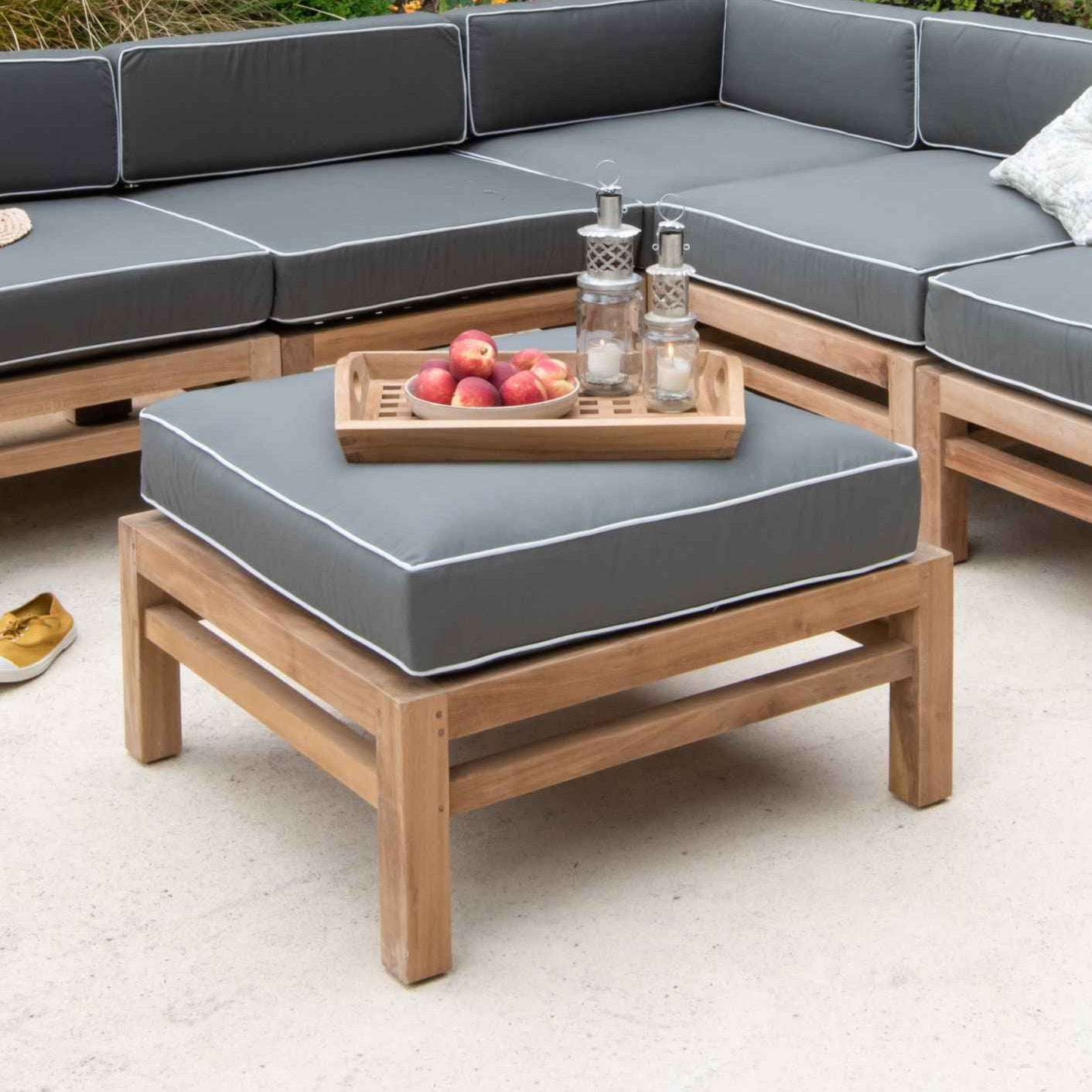 Teak outdoor sofa and ottoman stool with green and white cushions. The stool is holding a teak tray of fruit and lanterns. The floor is a stone patio 