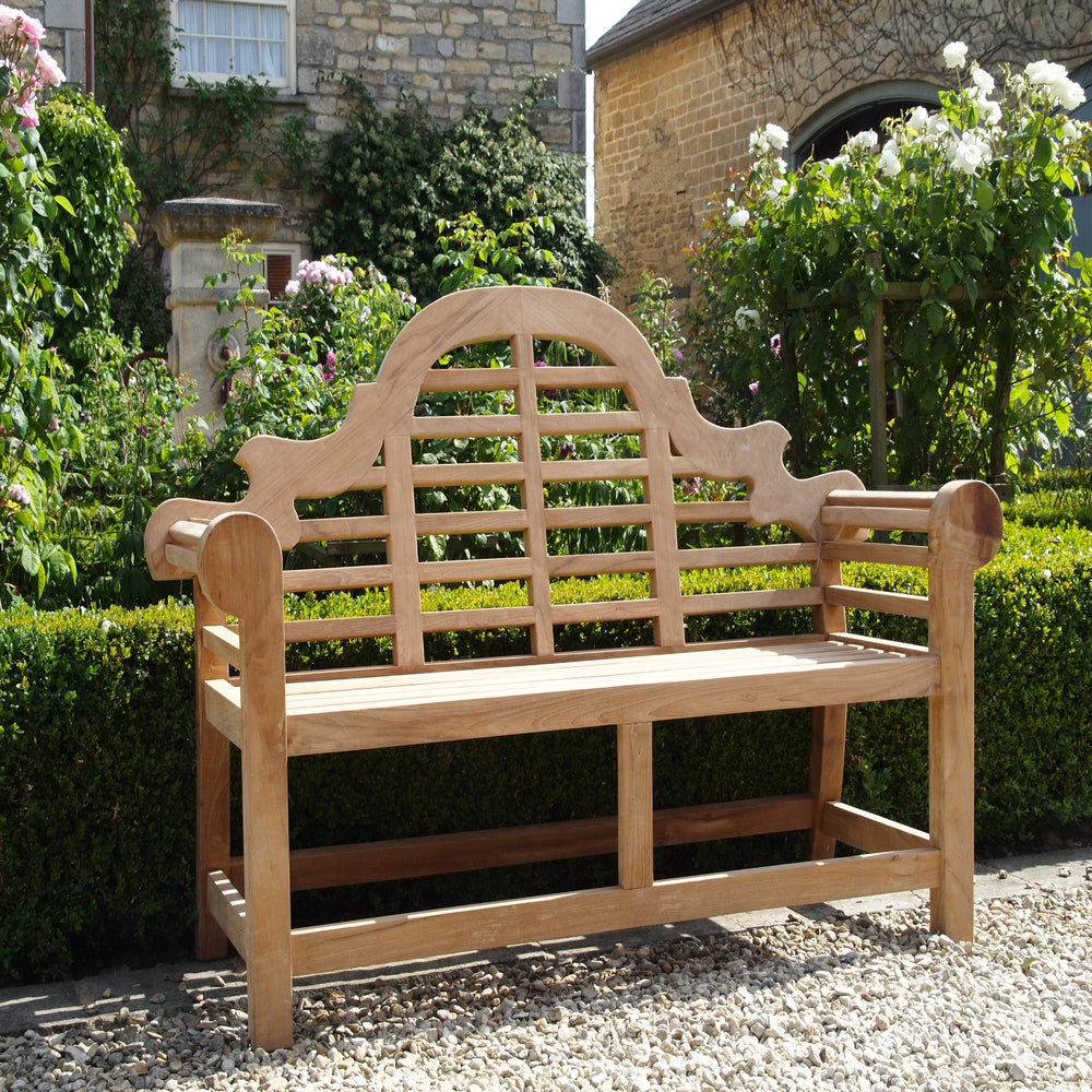 
                  
                    Teak bench with ornate, curved backrest and armrests and slatted seat, on gravel path with neat hedges and rose bushes behind. 
                  
                