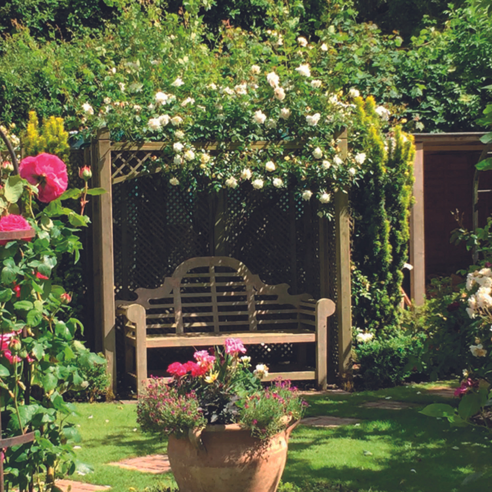 
                  
                    An ornate Lutyens-inspired teak garden bench with a intricately curved cutout back and rounded scroll-shaped arms, situated in a garden setting with floral backgrounds.
                  
                