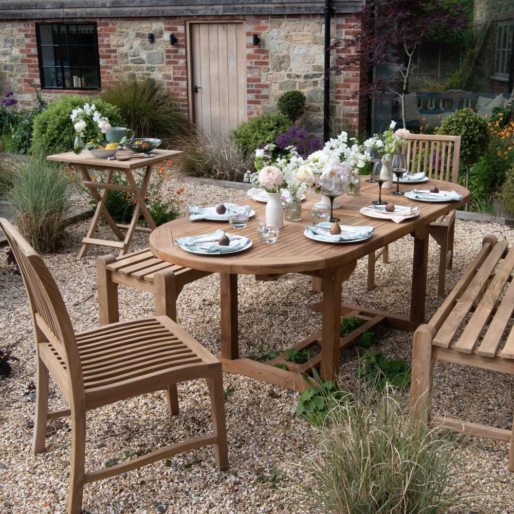 
                  
                    Teak benches without back or armrests, and teak dining chairs, around teak extending, oval dining table, set with plates, glasses and flowers, with brick and wood building behind. 
                  
                