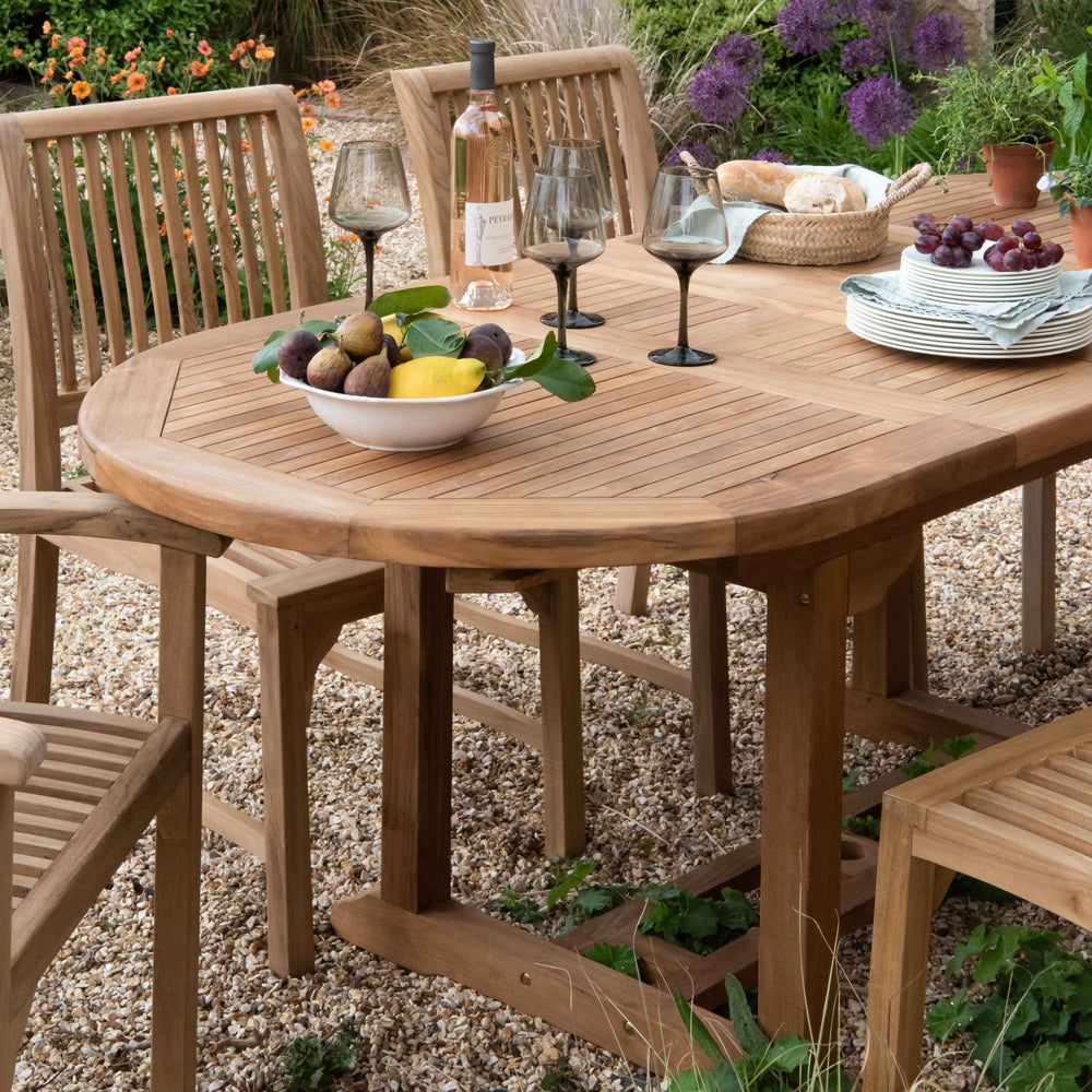 
                  
                    Teak Oval extending table with wine and glasses, fruit bowl and plates, with teak dining chairs set around, on gravel patio. 
                  
                