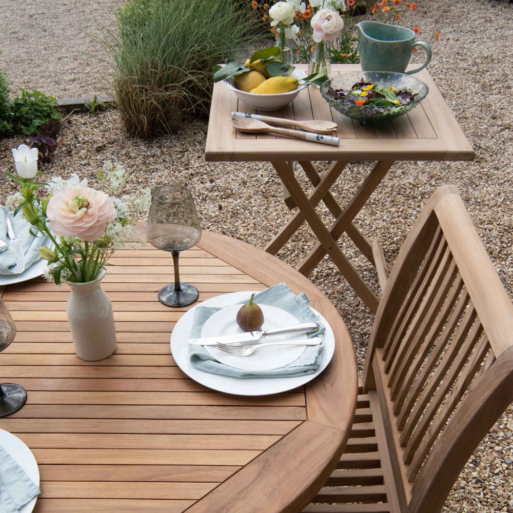 
                  
                    Teak Oval extending dining table with glasses and place settings, with small square teak table to the side, with salad bowl and servers, on gravel patio
                  
                