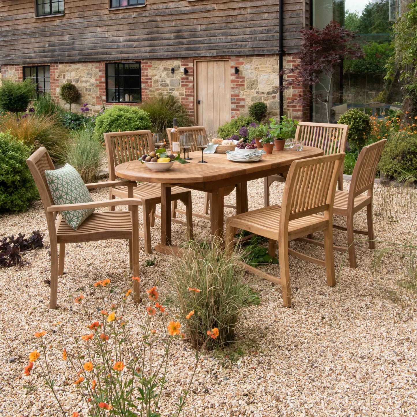 Teak oval extending dining table with wine glasses, fruit bowl and plants, and teak dining chairs set around, in front of wood and brick building, on gravel patio