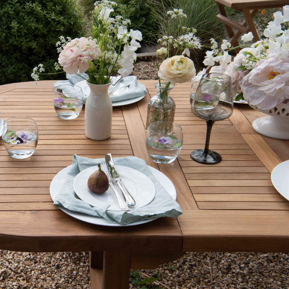 
                  
                    Teak oval extending table with wine glasses, place settings and vases of flowers, on gravel patio 
                  
                