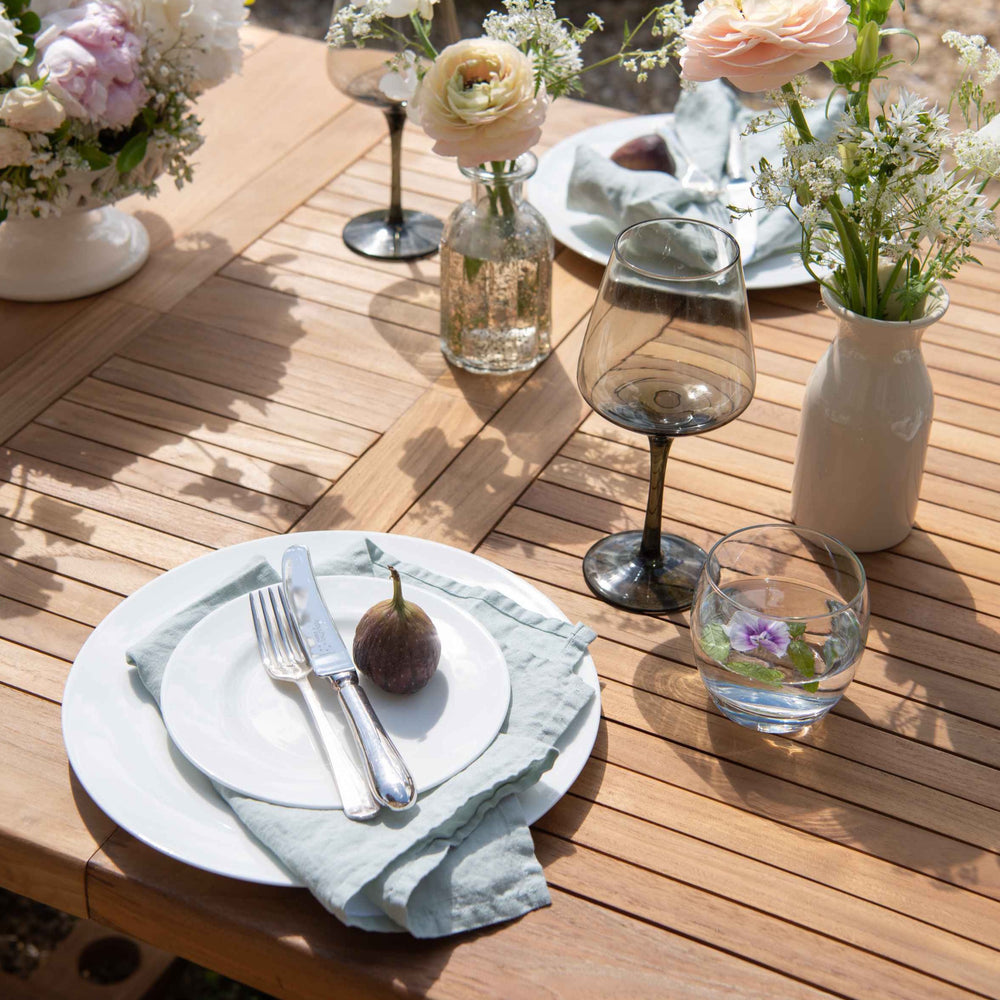 
                  
                    Close up of teak oval table with slatted tabletop, set with place settings, glasses and flowers
                  
                