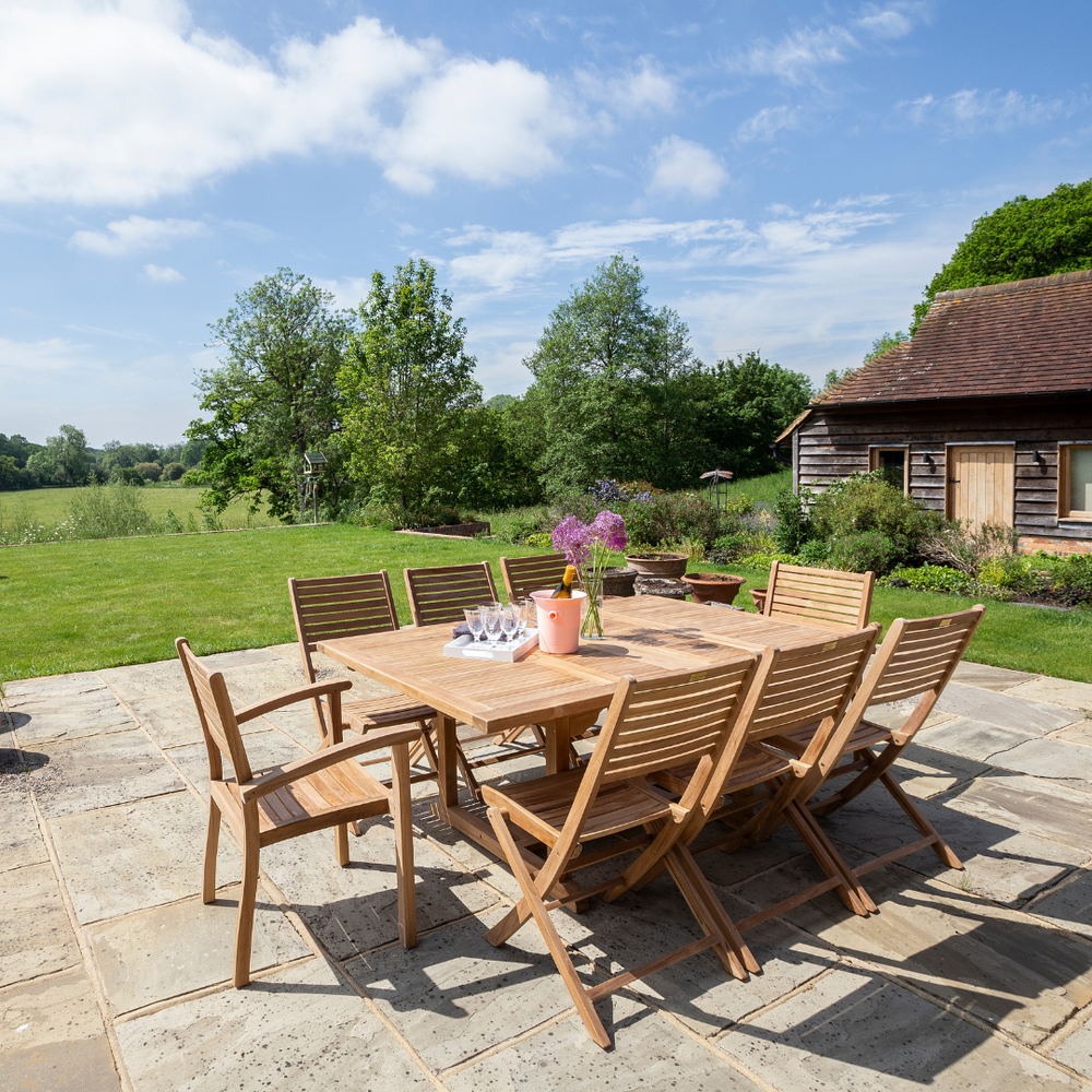 
                  
                    Extending teak rectangular table with glasses, flowers and wine, six teak folding chairs and two teak chairs with armrests, on stone patio with garden and buildings in background. 
                  
                