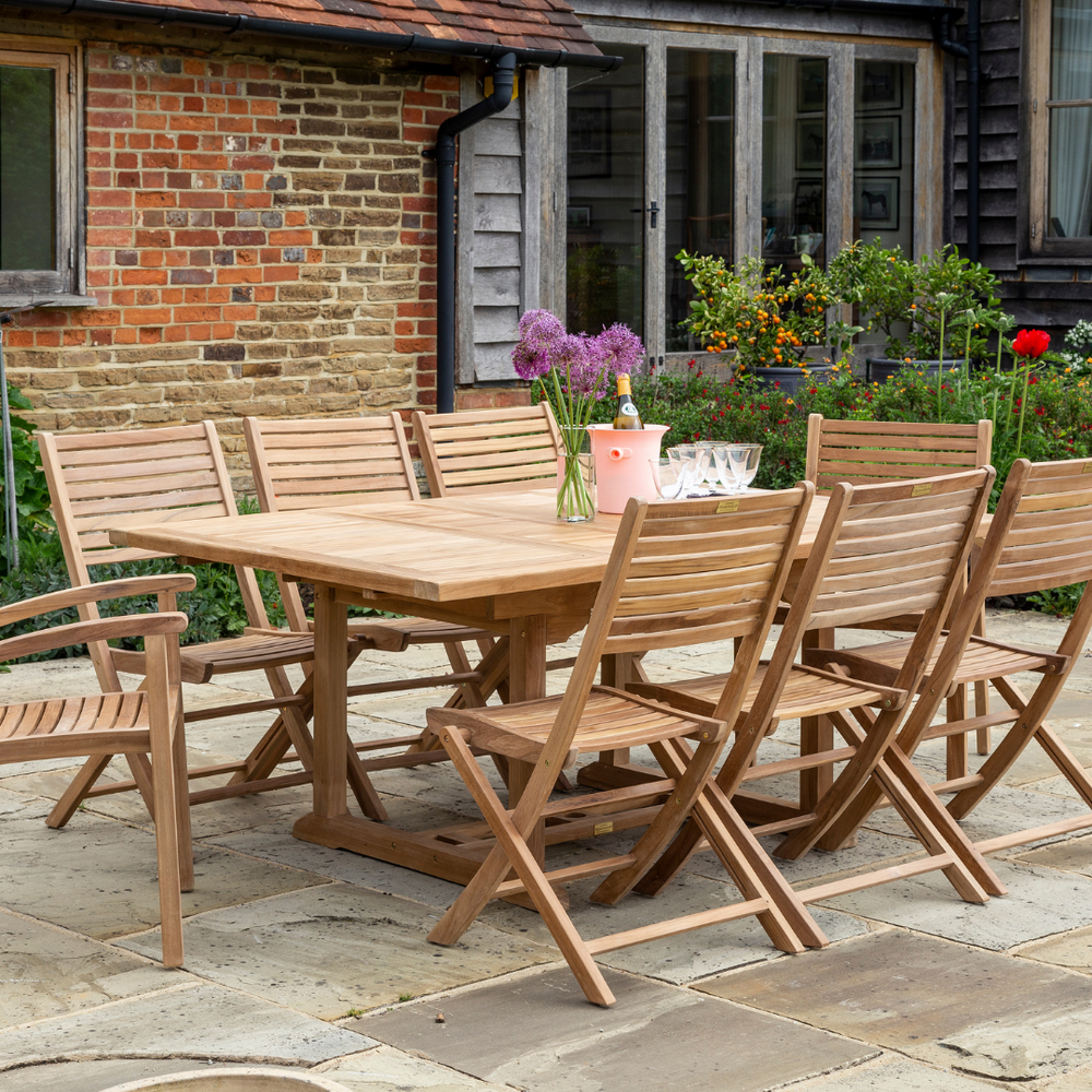 
                  
                    Teak rectangular dining table with wine, flowers and glasses with 6 teak folding dining chairs set around, in front of red brick and wooden building. 
                  
                