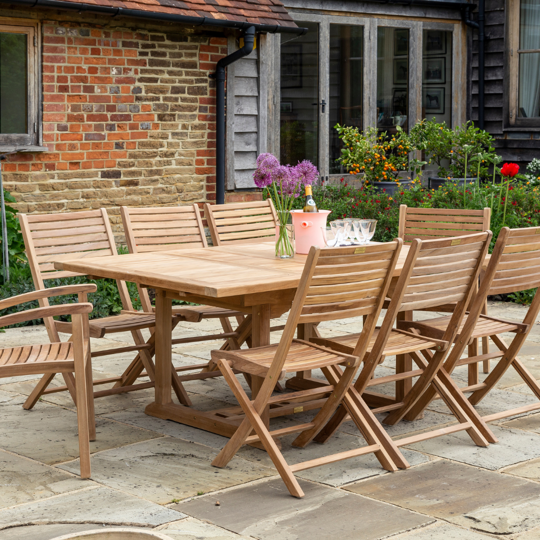 Teak rectangular dining table with wine, flowers and glasses with 6 teak folding dining chairs set around, in front of red brick and wooden building. 