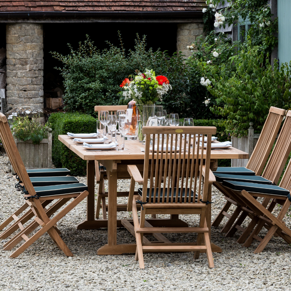 
                  
                    Teak rectangular extending dining table laid with glasses and place settings, with six folding teak chairs with green seat cushions, and two teak carver chairs with armrests and cushions.  In the background are plants and stone pillars. 
                  
                