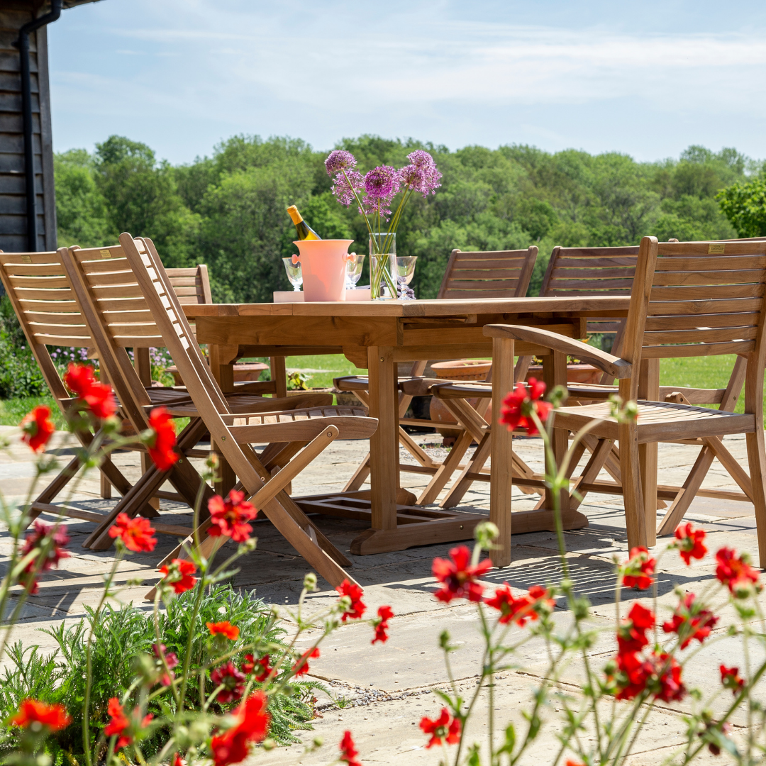 Rectangular extending teak dining table with teak folding and carver chairs around, red flowers in the foreground and wine, flowers and glasses on the table. 