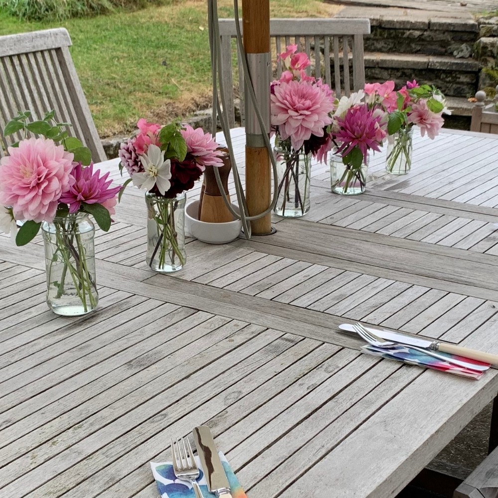 
                  
                    Weathered rectangular teak dining table with teak chairs, parasol and glass vases of pink flowers
                  
                