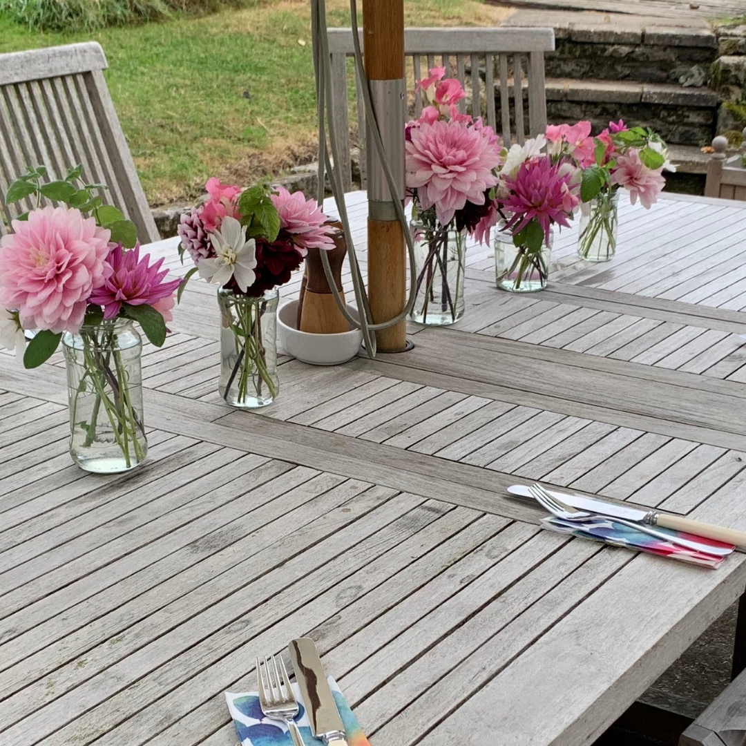 Weathered rectangular teak dining table with teak chairs, parasol and glass vases of pink flowers
