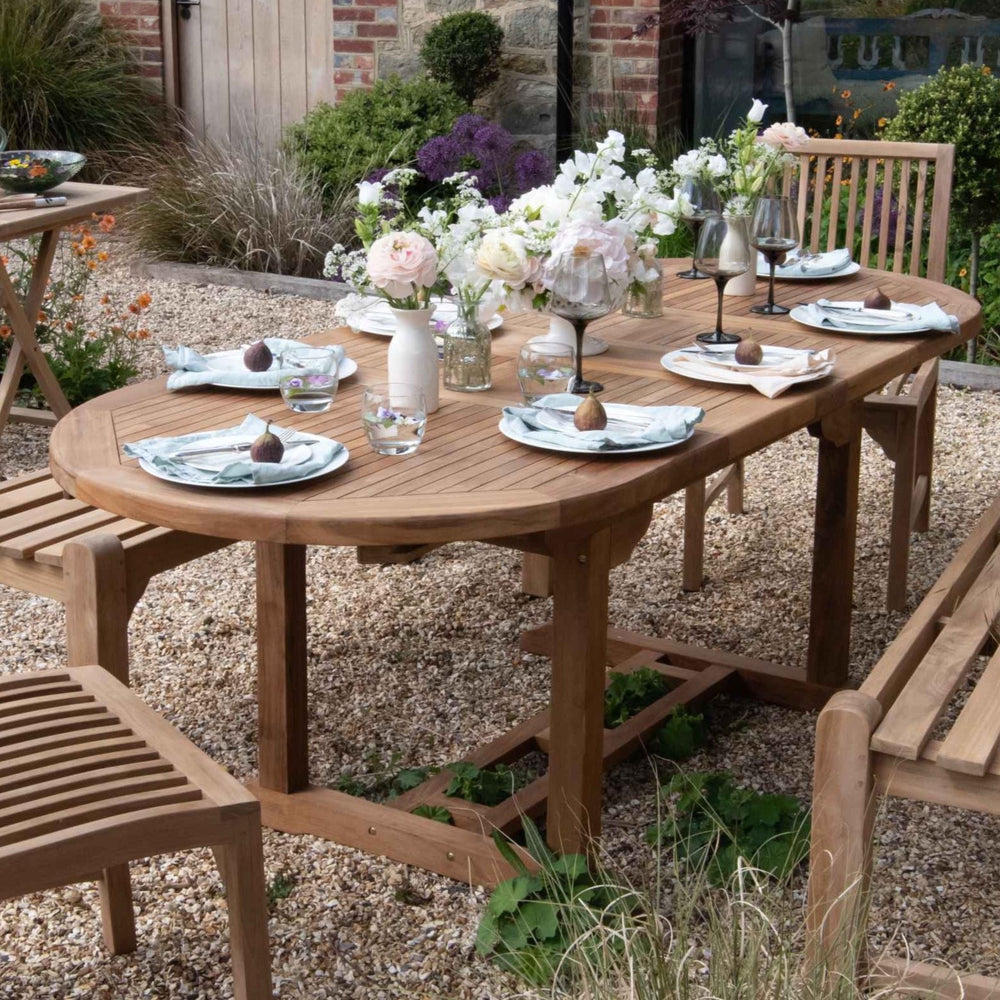 An oval teak outdoor dining table set with plates, glasses, and a floral centerpiece, on a gravel surface with teak chairs and. benches in the background.