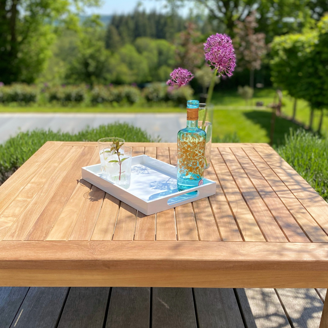 Teak square coffee table, low to the ground, on wooden deck with garden and tennis court in background. On the table is a white tray with glass, flowers and blue bottle.