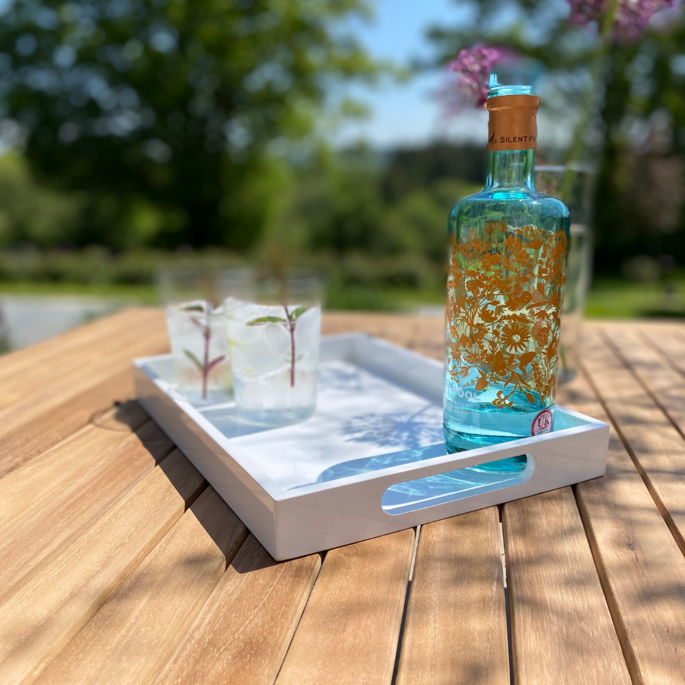 
                  
                    Close up of table top of teak square table top with white tray of glasses and bottle and garden in background.  
                  
                