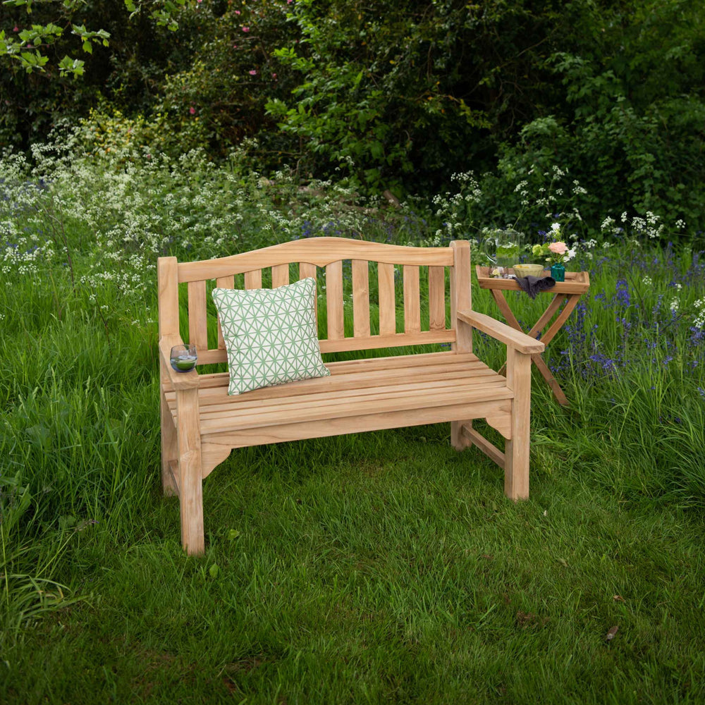 
                  
                    Teak Bench with curved backrest, straight arms, next to small square folding teak table, with green patterned scatter cushion, in garden setting 
                  
                