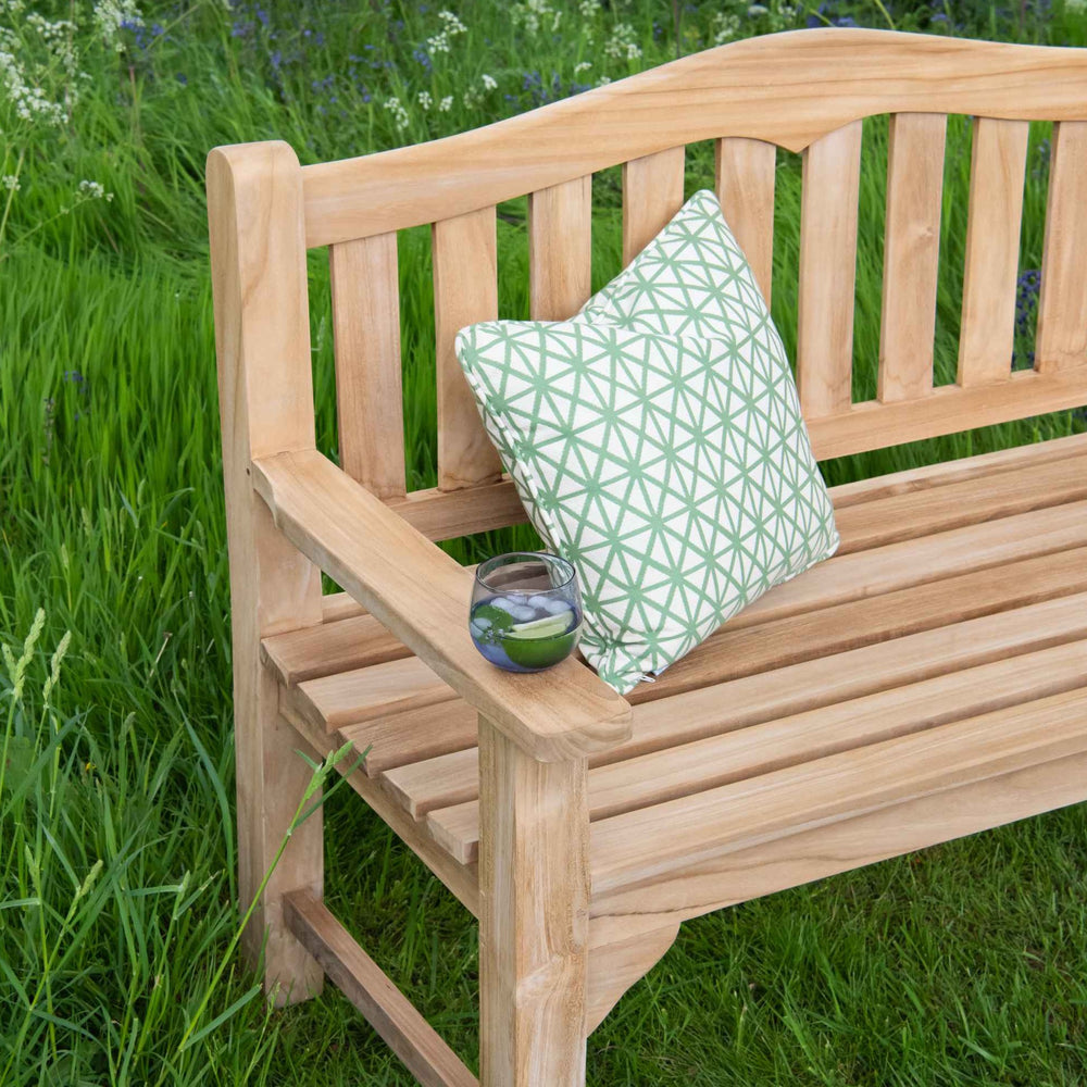 
                  
                    Teak Bench with curved backrest and straight arms. On the arm is a glass next to a green patterned cushion. The bench is sitting on long grass. 
                  
                