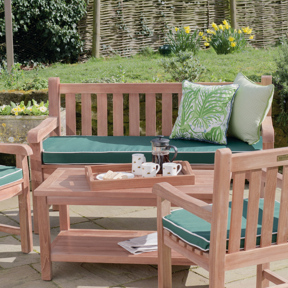 
                  
                    Teak outdoor bench and teak chairs with armrests, all with green seat cushions, around teak rectangular coffee table with tray of coffee and cups, in garden setting. 
                  
                