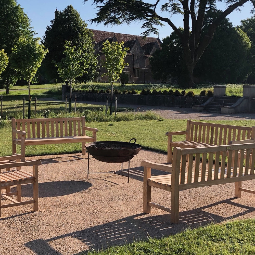 
                  
                    Four teak benches with square, slatted backrests and arms, set around a metal fire pit, on a gravel patio. Behind is lawn and trees.  
                  
                
